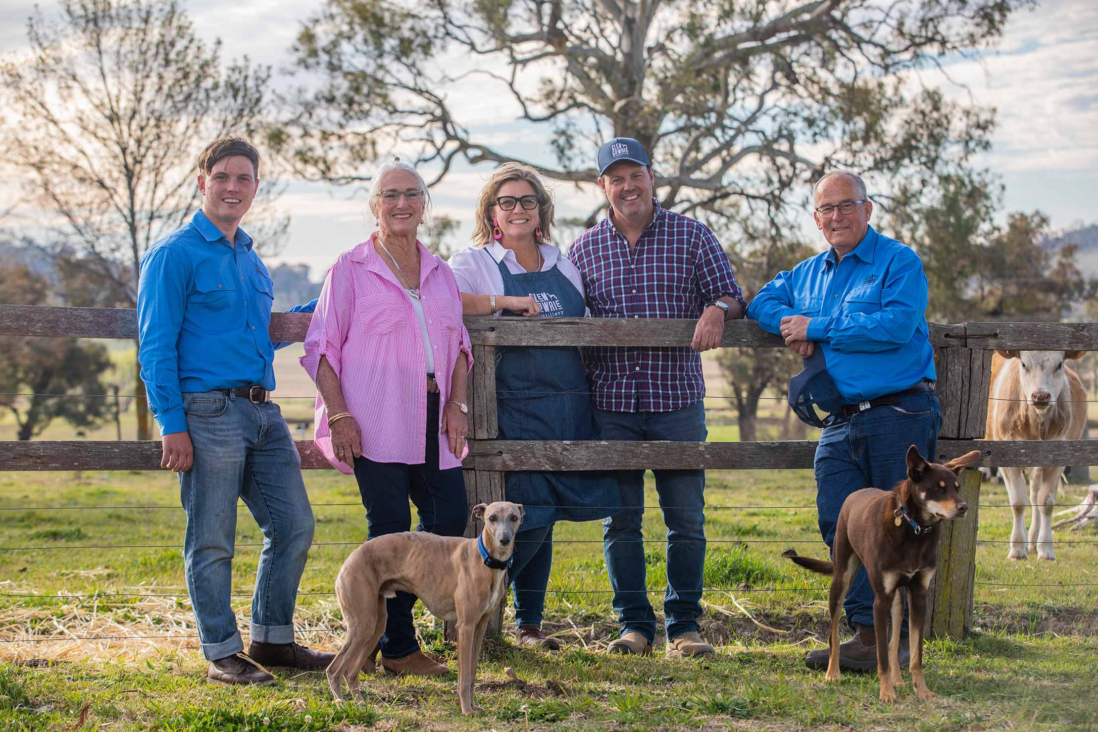 Five people who make stout and two dogs pose by a wooden fence in a green field, with trees and cows in the background.