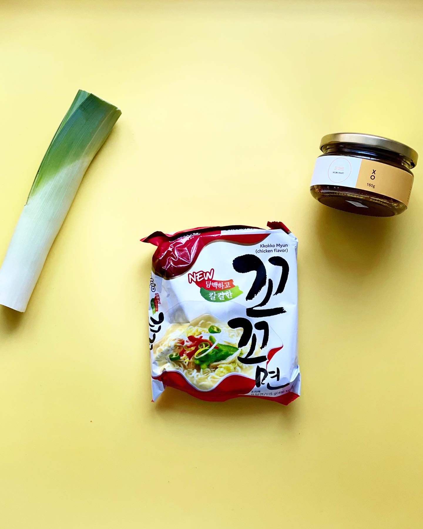 A pack of Korean noodles with a jar of XO sauce and a leeks on a yellow background.
