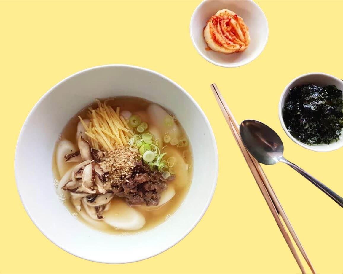 Bowl of noodle soup with beef, mushrooms, ginger, and green onions, accompanied by kimchi and seaweed.