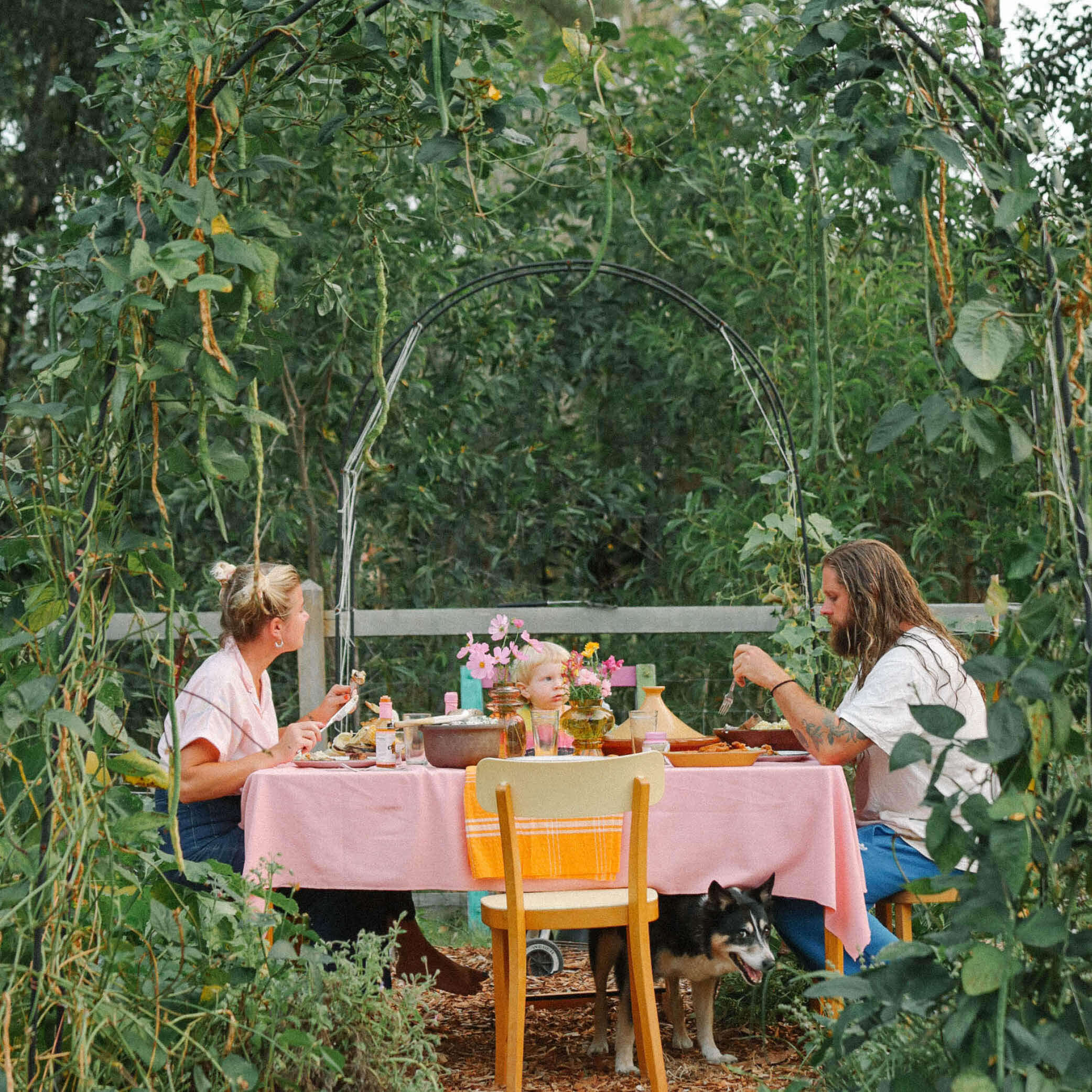 Family enjoying a meal outdoors at a garden table, surrounded by greenery and a playful dog.