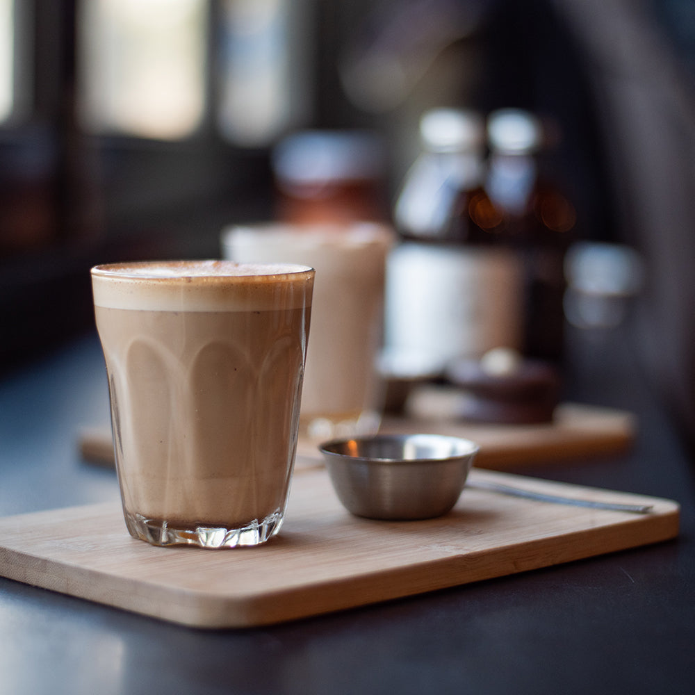 Glass of creamy chai latte on a wooden board with spices and bottles in the background.
