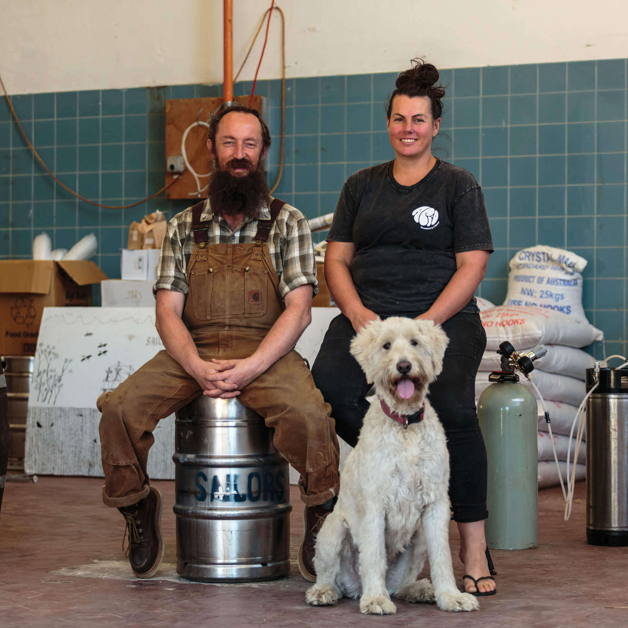 Two people and a dog in a warehouse, showcasing a friendly atmosphere, with brewing supplies in the background.