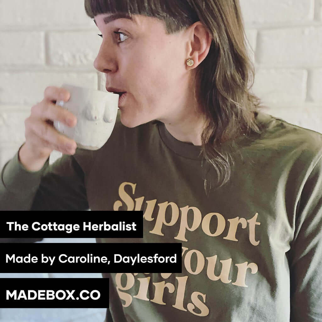 Woman wearing a 'Support Your Girls' shirt, enjoying a beverage, promoting The Cottage Herbalist and local artisans.