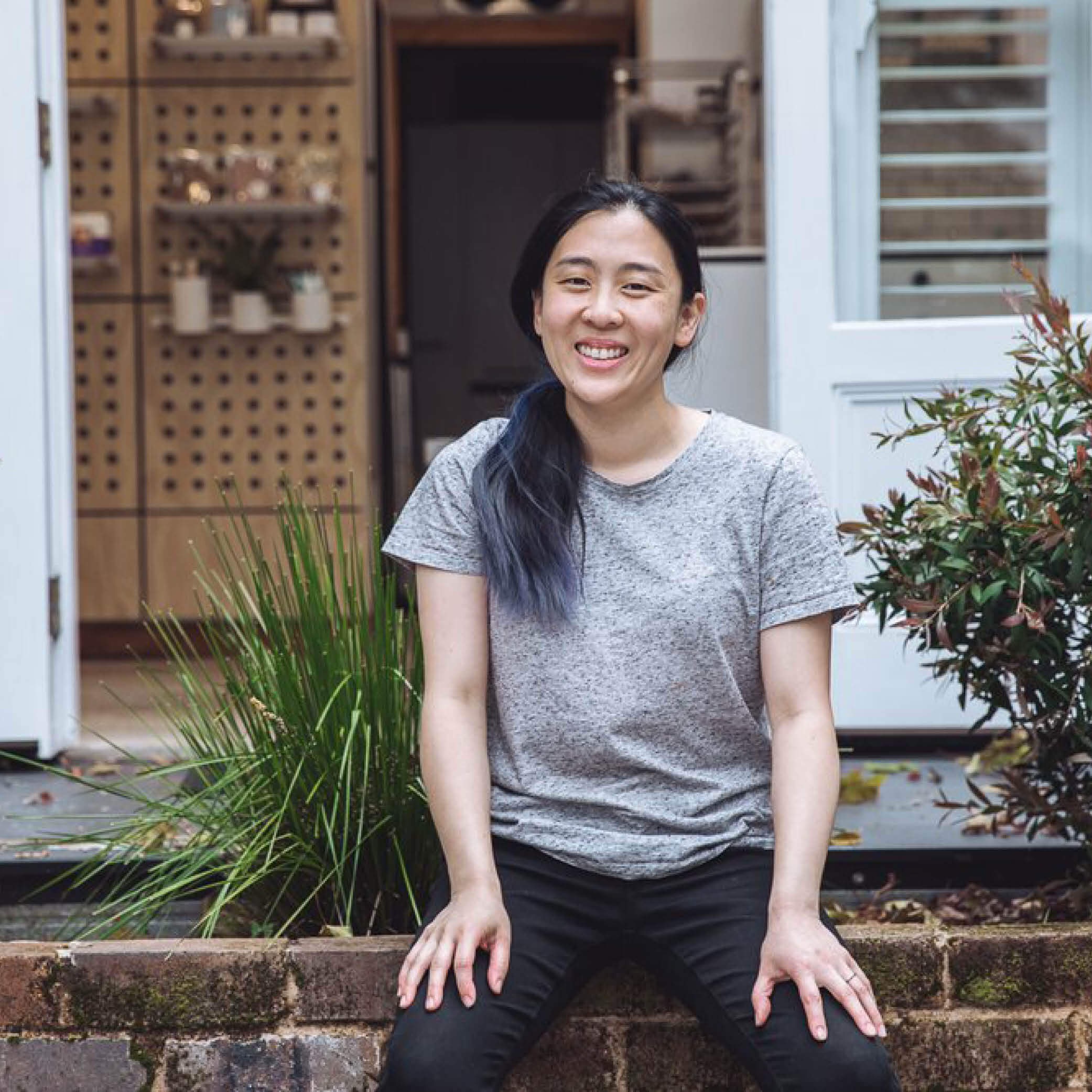 Smiling person sitting on steps in casual attire with greenery in the background, radiating a friendly and relaxed vibe.