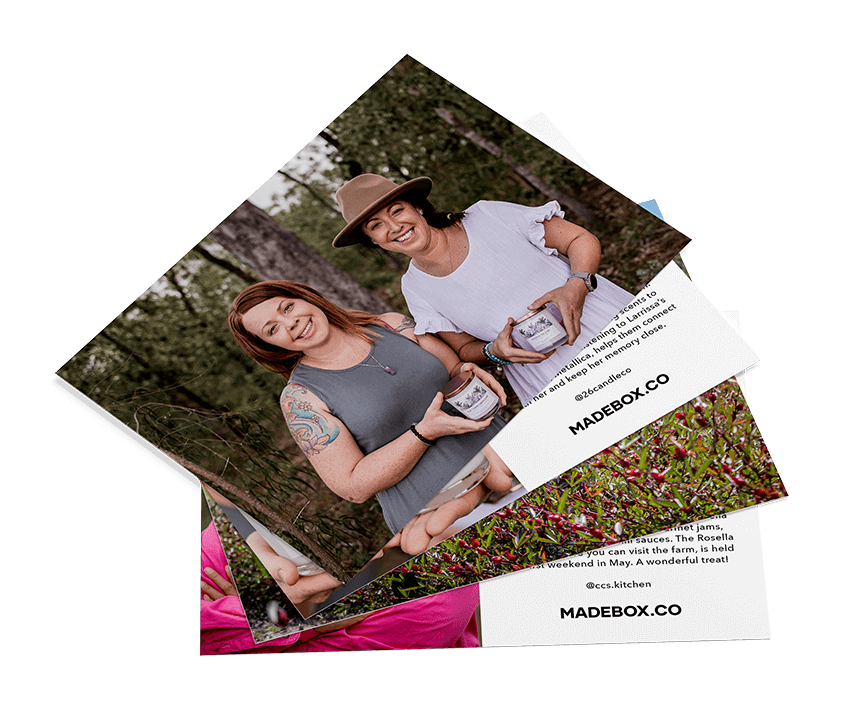 Two women smiling while holding gourmet BBQ boxes in a scenic outdoor setting surrounded by nature.