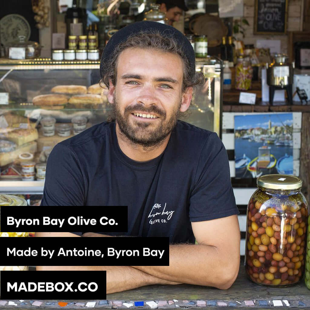 Smiling man at Byron Bay Olive Co. with jars of olives, promoting local Australian food.