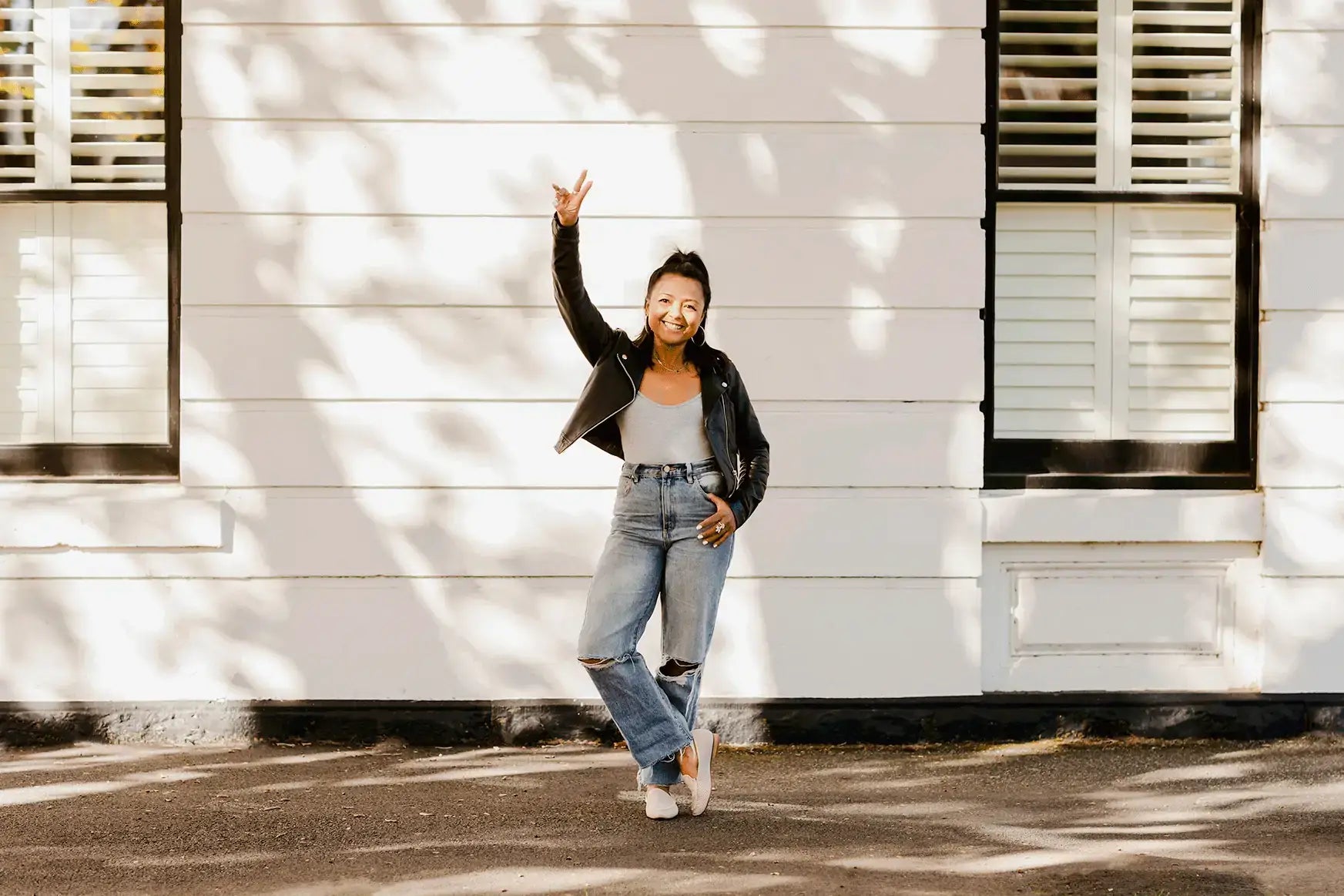 Smiling woman in a leather jacket and jeans poses confidently with a peace sign against a bright wall.