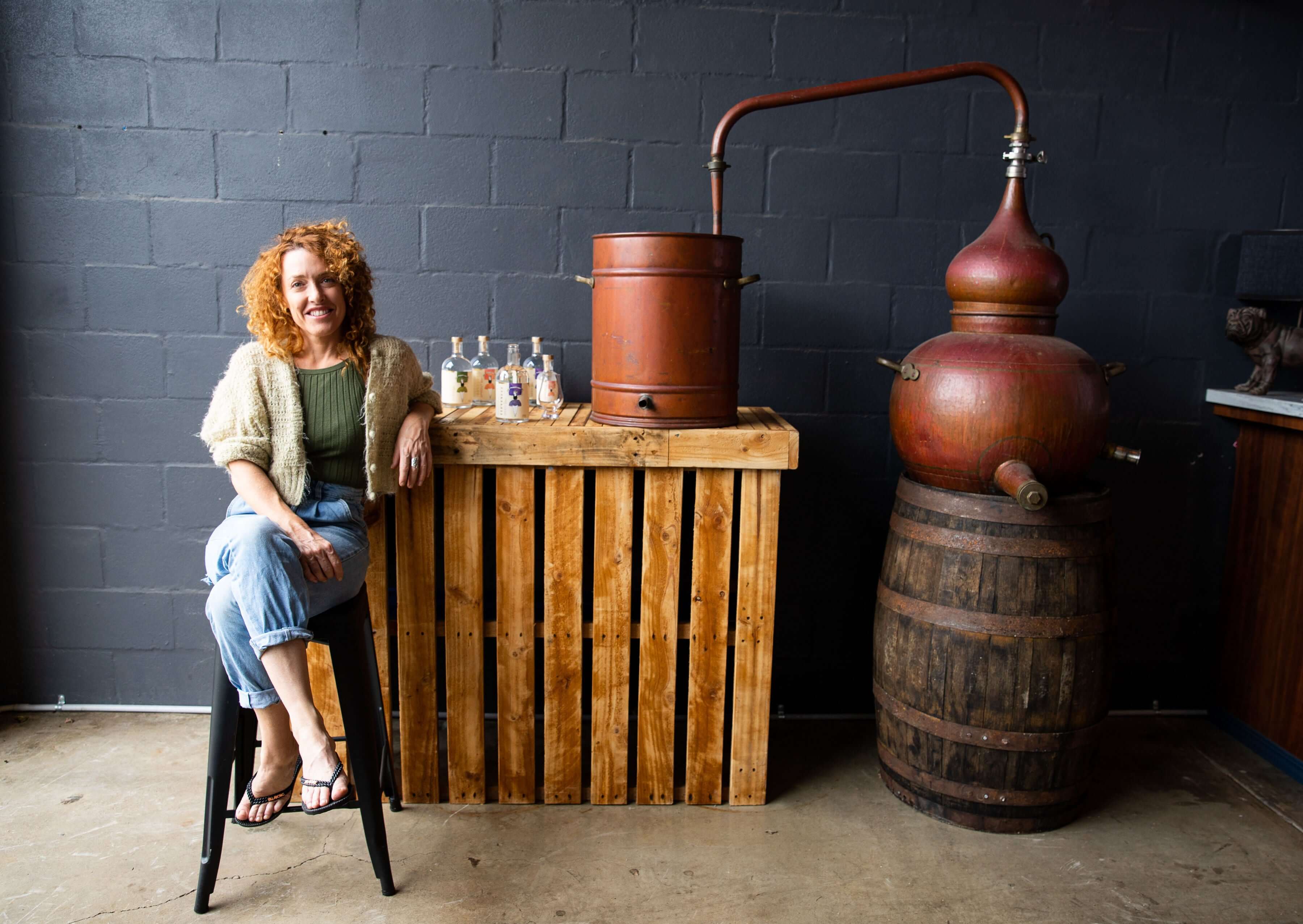 Woman sitting on a stool beside a wooden table and vintage distillation equipment in a modern distillery.