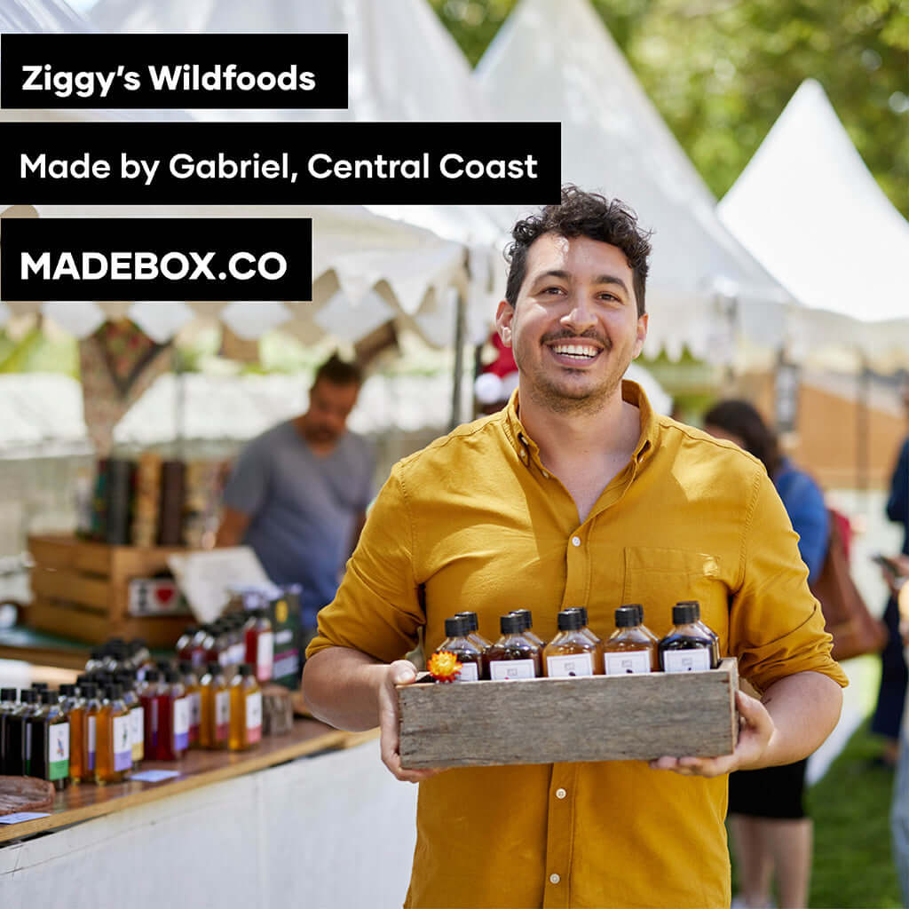 Man holding a wooden box of Ziggy's Wildfoods products at a market in Central Coast, smiling in a yellow shirt.